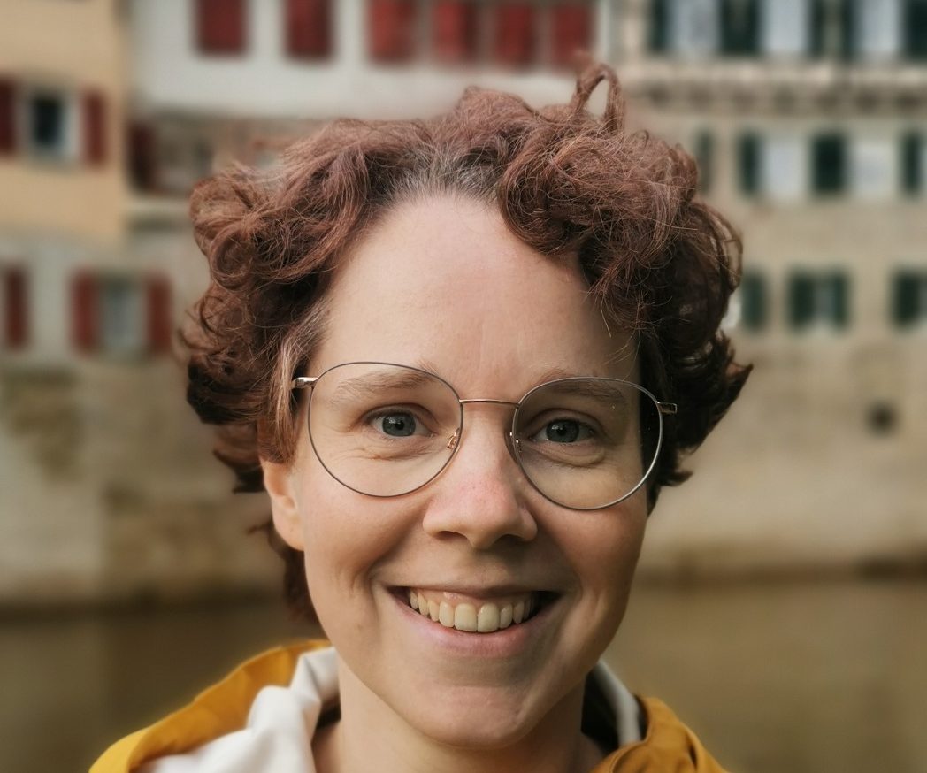 Profil einer lächelnden Frau mit kurz geschnittenen Locken vor einer historischen Häuserfassade